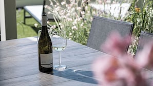 A bottle of wine and a glass on a wooden table outdoors with a garden in the background.