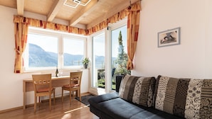A cozy living room with a wooden ceiling, a dark gray sofa with patterned cushions, a dining area with a wooden table and chairs, and large windows offering a mountain view.