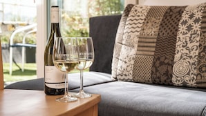 A bottle of wine and two glasses on a wooden table in a room with a patterned cushion and a view of greenery outside.