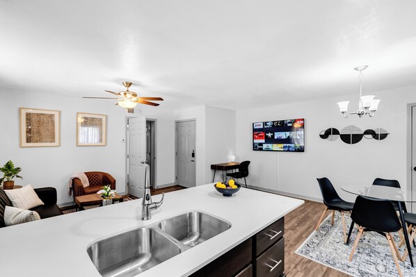 A modern kitchen with a large island, a dining area with a table and chairs, and a living room with a sofa and a TV mounted on the wall.