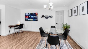 Un comedor moderno con una mesa de vidrio, sillas negras, un escritorio de madera y un televisor montado que muestra varios canales.