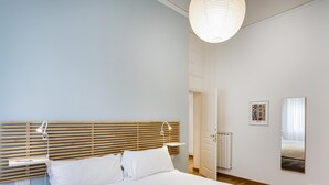 A bedroom with a wooden slatted headboard, a white bed with pillows, a bedside table with a lamp, a radiator, and a framed picture on the wall. - Light Apartment in Albaro (Genoa)