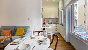 A dining area with a round table set for four, a sofa with cushions, and a view of the kitchen and balcony. - Light Apartment in Albaro (Genoa)