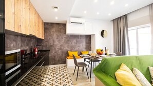 A modern kitchen with a microwave, stove, and wooden cabinets. There is a dining area with a table, chairs, and a green sofa with yellow pillows.