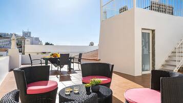 Terraço no telhado com mobiliário de vime, uma mesa com um vaso de flores e vista para a paisagem urbana.