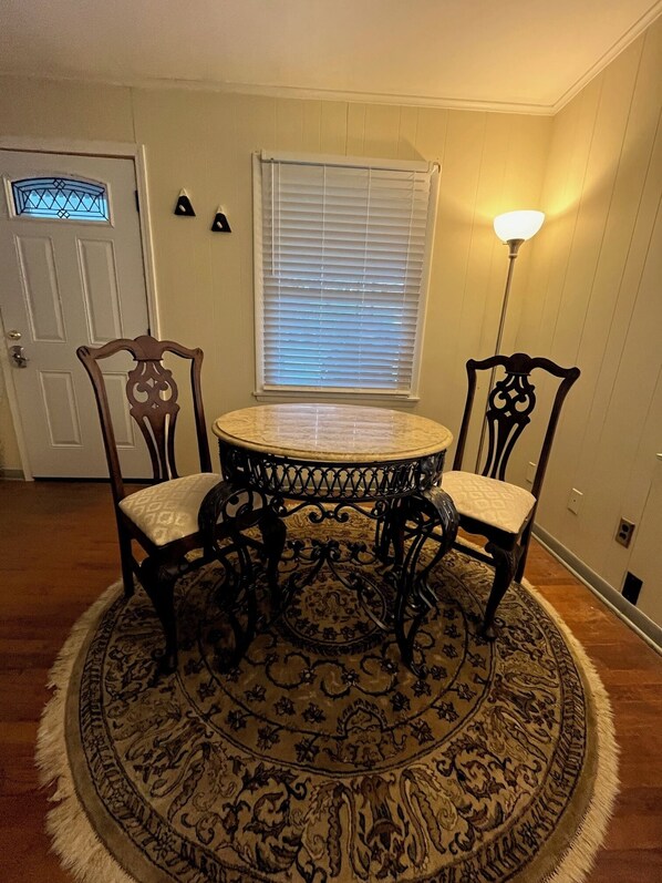 Un comedor con una mesa redonda, dos sillas, un tapete estampado, una ventana con persianas y una puerta.