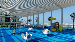 Een overdekt buitengebied met een blauw geruit vloerkleed, een speelconstructie voor kinderen en uitzicht op zee.