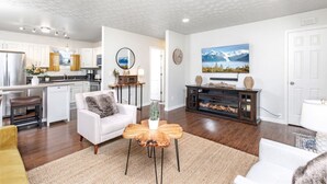A modern living room with a white sofa, a coffee table, and a fireplace. There is a kitchen in the background with white cabinets and a refrigerator.