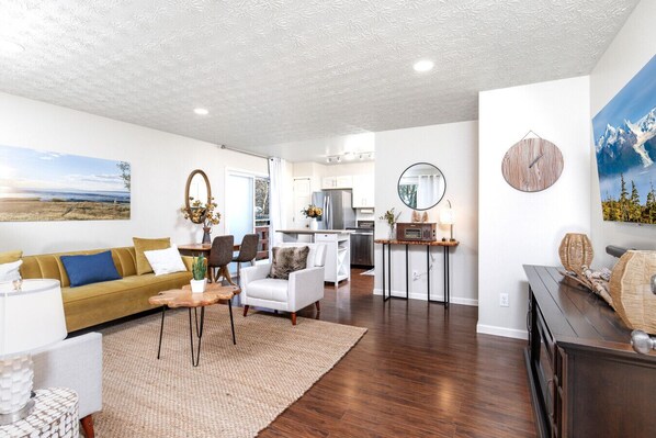 A modern living room with a sofa, armchair, and coffee table, a kitchen area, and a dining space.