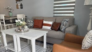 Un salón con un sofá gris, una mesa de centro blanca y un sillón de cuero marrón. Hay un jarrón con flores sobre la mesa de centro.