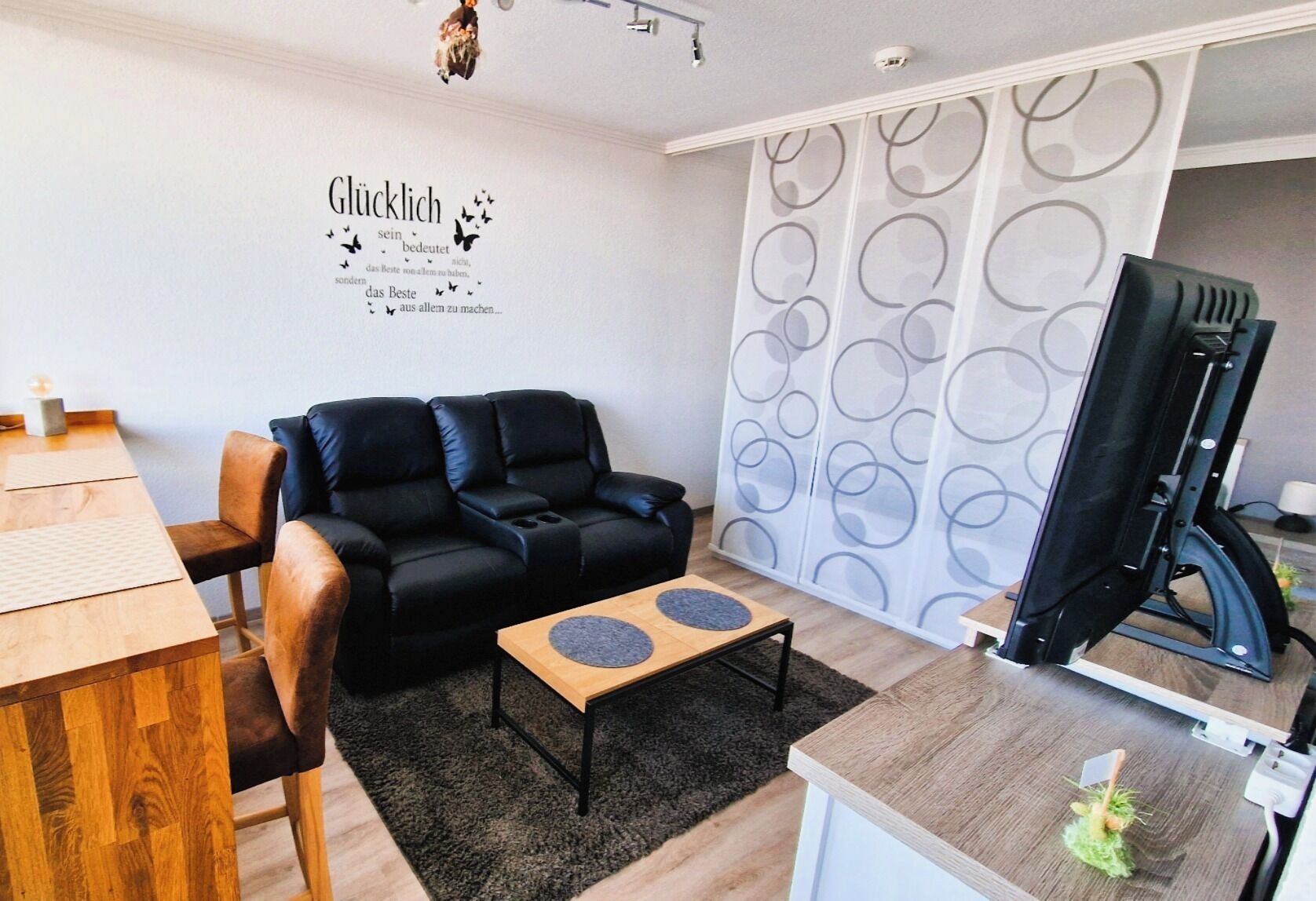 Ein modernes Wohnzimmer mit einem schwarzen Ledersofa, einem Holztisch und einer Wand mit einem Motivationsspruch.