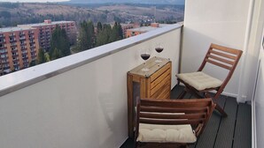 Un balcon avec une chaise en bois et une petite table, deux verres de vin rouge, et une vue sur des bâtiments et des collines.