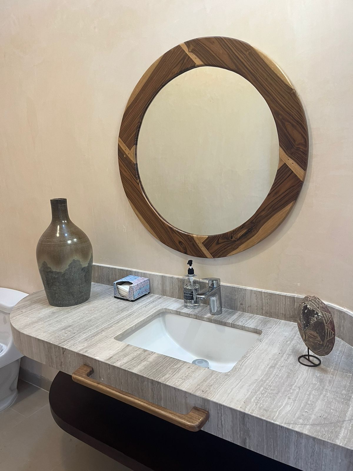 A bathroom with a round mirror, a sink, a vase, a soap dispenser, and a towel holder.