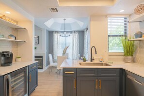 A modern kitchen with dark cabinetry, a white countertop, and a built-in oven. There is a window with blinds and a potted plant on the counter.