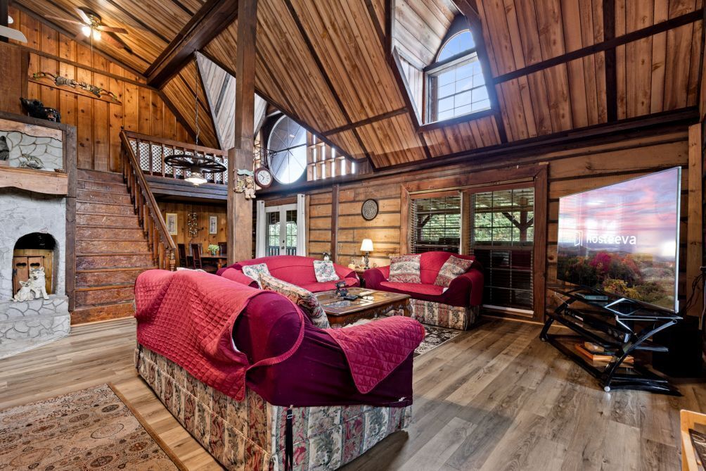 A cozy log cabin interior with a fireplace, a sofa set, and a large window overlooking a scenic landscape.