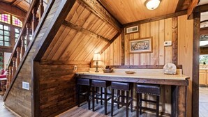 Un intérieur rustique en bois, avec un plafond en pente, un coin bar doté de tabourets et un tableau encadré au mur.