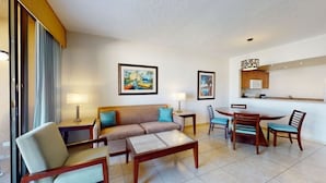 Une chambre d’hôtel comprenant un canapé, deux fauteuils, une table basse, une table à manger avec quatre chaises, un coin cuisine avec un four à micro-ondes et deux tableaux encadrés au mur.
