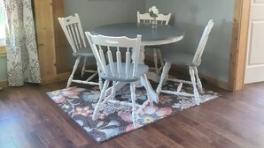 Comedor con una mesa redonda, cuatro sillas, un tapete floral, un ventilador de techo y una pared con una pintura de flores y símbolos de utensilios.