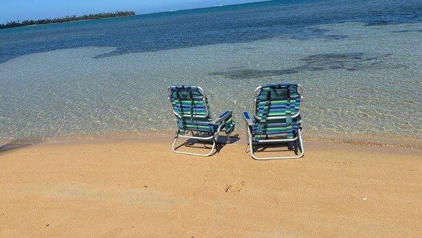 Dos sillas de playa sobre arena, con agua cristalina y una costa lejana.