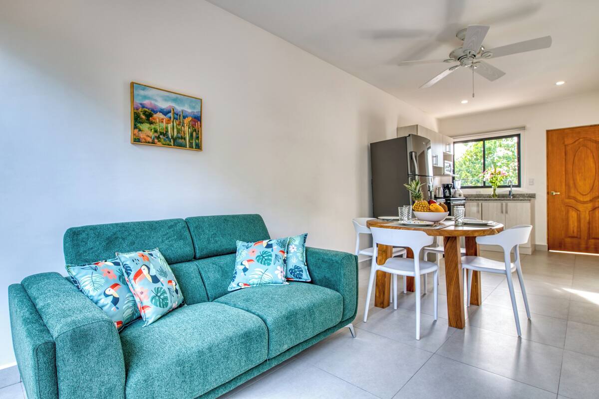 A modern living room with a teal sofa, white chairs, a wooden dining table, and a ceiling fan.