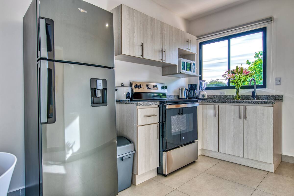 A modern kitchen with stainless steel appliances, light wood cabinets, and a large window providing natural light.