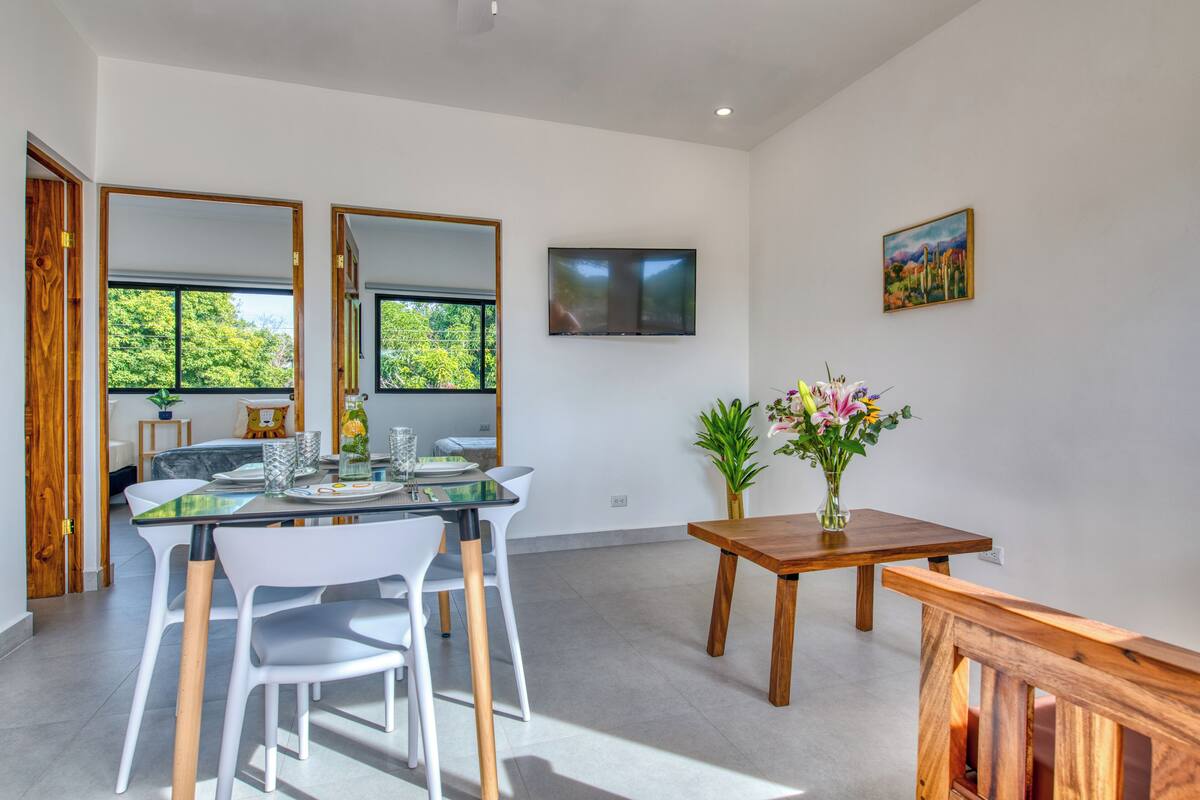 A modern dining area with a glass table, white chairs, a wooden side table with a vase of flowers, and a TV mounted on the wall.