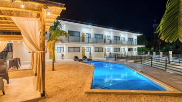 A hotel with a swimming pool, outdoor seating, and a covered area with a palm tree.