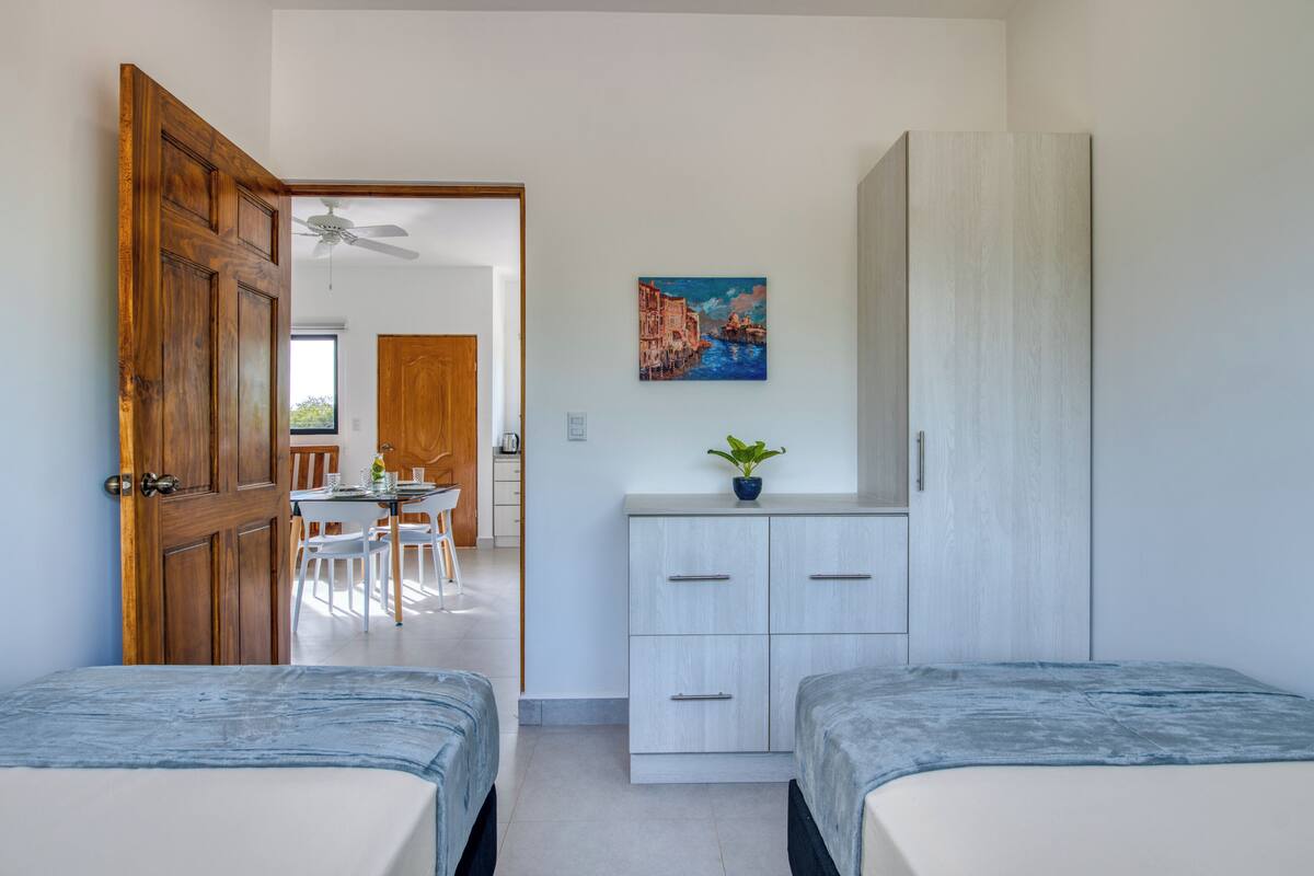 A bedroom with two beds, a wooden door, a desk with chairs, and a painting on the wall.