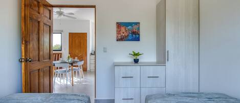 A bedroom with two beds, a wooden door, a desk with chairs, and a painting on the wall.