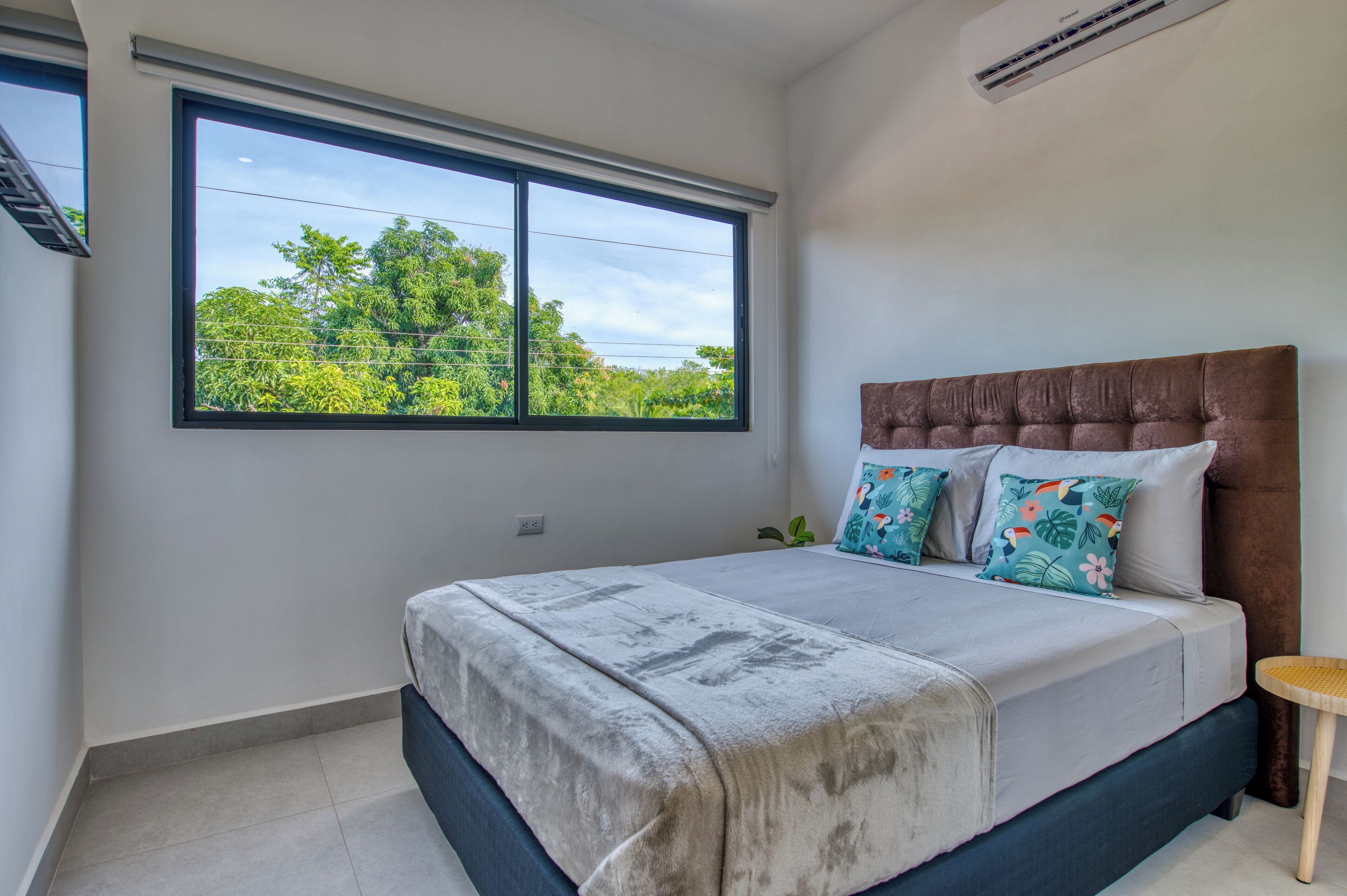 A bedroom with a large window, a bed with a brown headboard, and a small wooden stool.