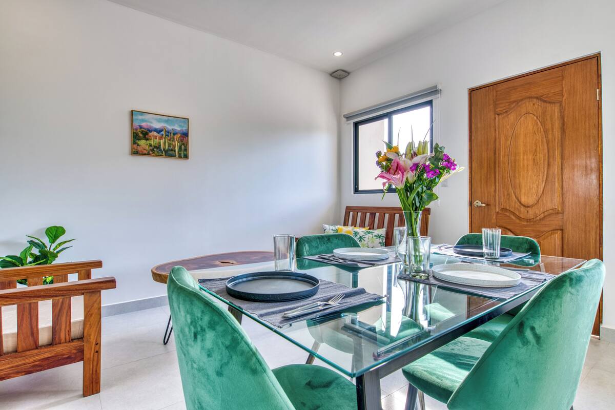 A dining area with a glass table, green velvet chairs, and a wooden door.