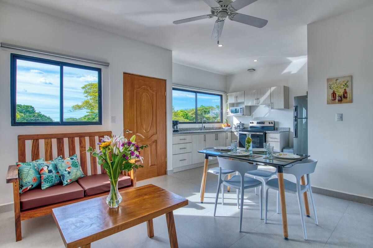 A modern kitchen with white cabinets, a dining table, and chairs. There is a wooden bench with cushions and a vase of flowers.