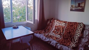 A room with a window, a table, a chair, a sofa with a patterned blanket, and an air conditioning unit.