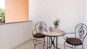 En lille udendørs terrasse med et rundt bord, to stole, en potteplante og flisegulv.