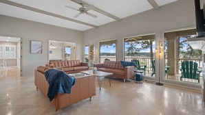 A spacious living room with a large sectional sofa, a coffee table, and a ceiling fan. There are large windows offering a view of the outdoors. - Beautiful, Private Lake House with Double Dock (Hot Springs)
