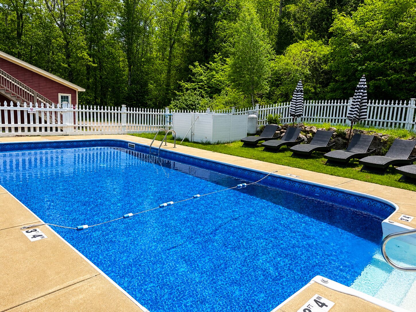 Ein rechteckiger Swimmingpool mit weißer Liegefläche, umgeben von einem weißen Ziergartenzaun, und ein kleines Gebäude zur Linke.