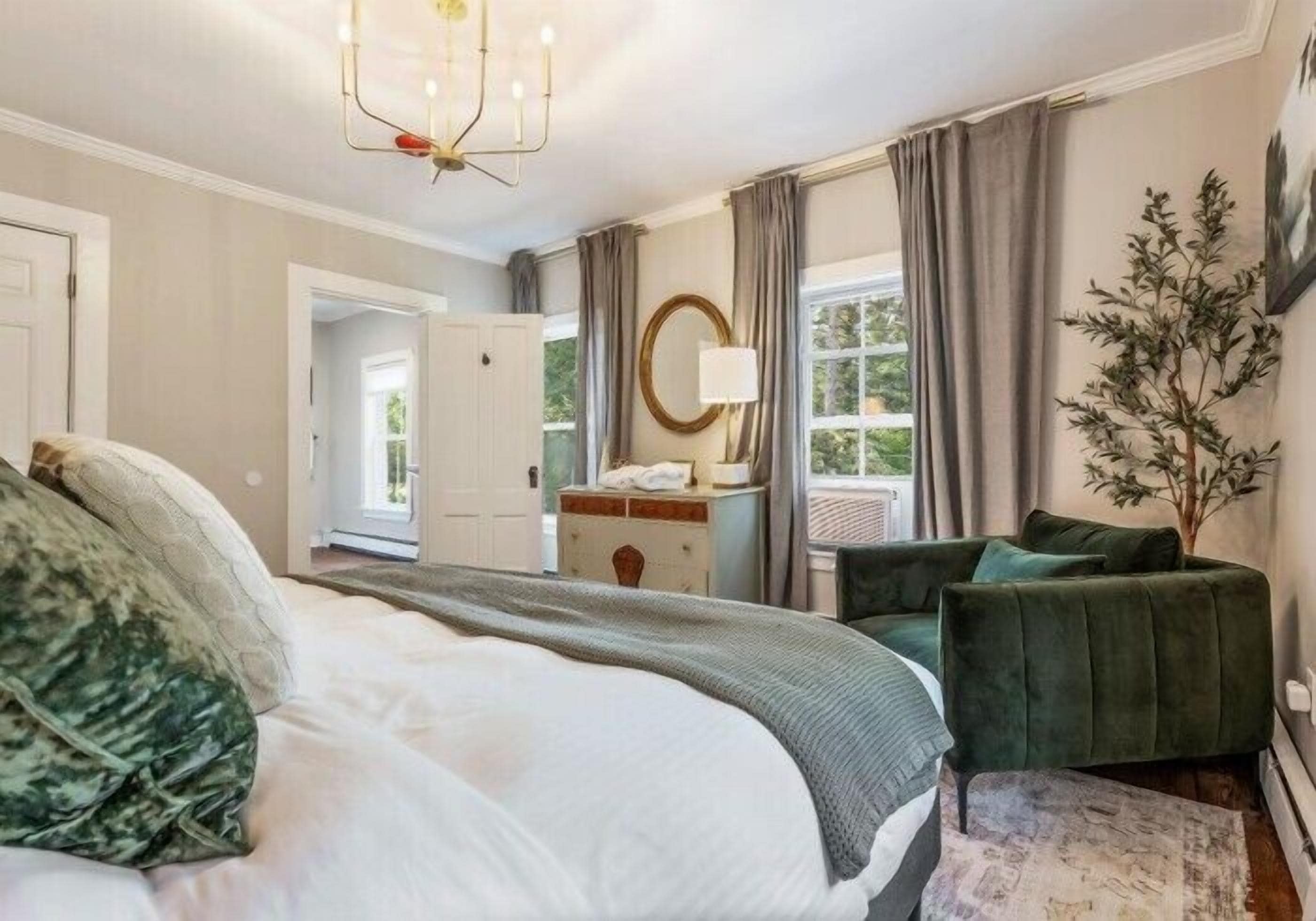 A bedroom with a bed, a green armchair, a dresser, a mirror, and a window with curtains.