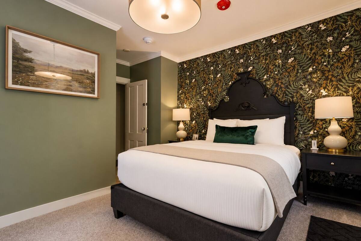 A bedroom with a bed, two bedside tables, a nightstand, a door, a framed picture on the wall, and a ceiling light fixture.