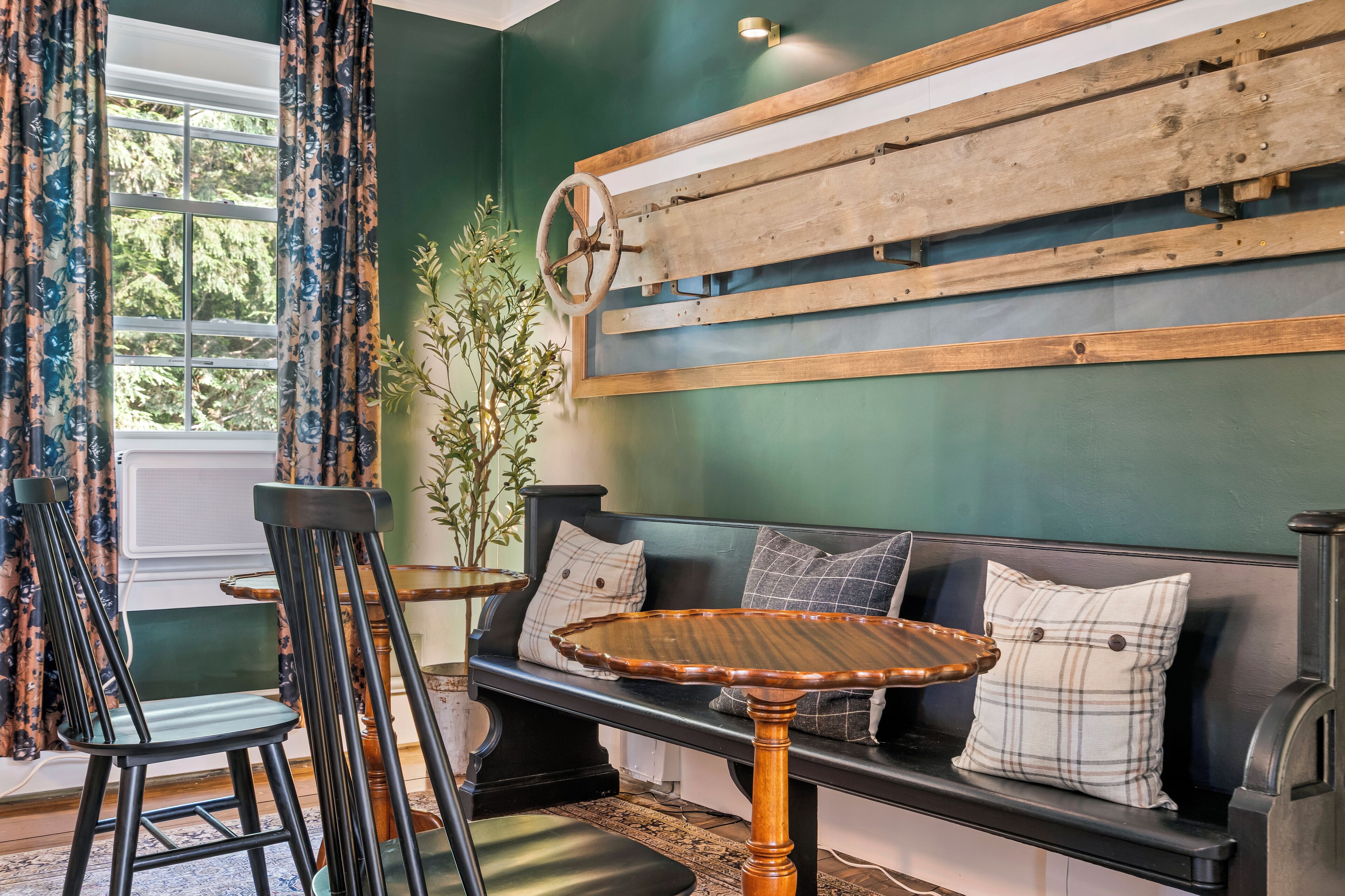 A room with a dark green wall, a wooden bench with cushions, a round wooden table, and two chairs.
