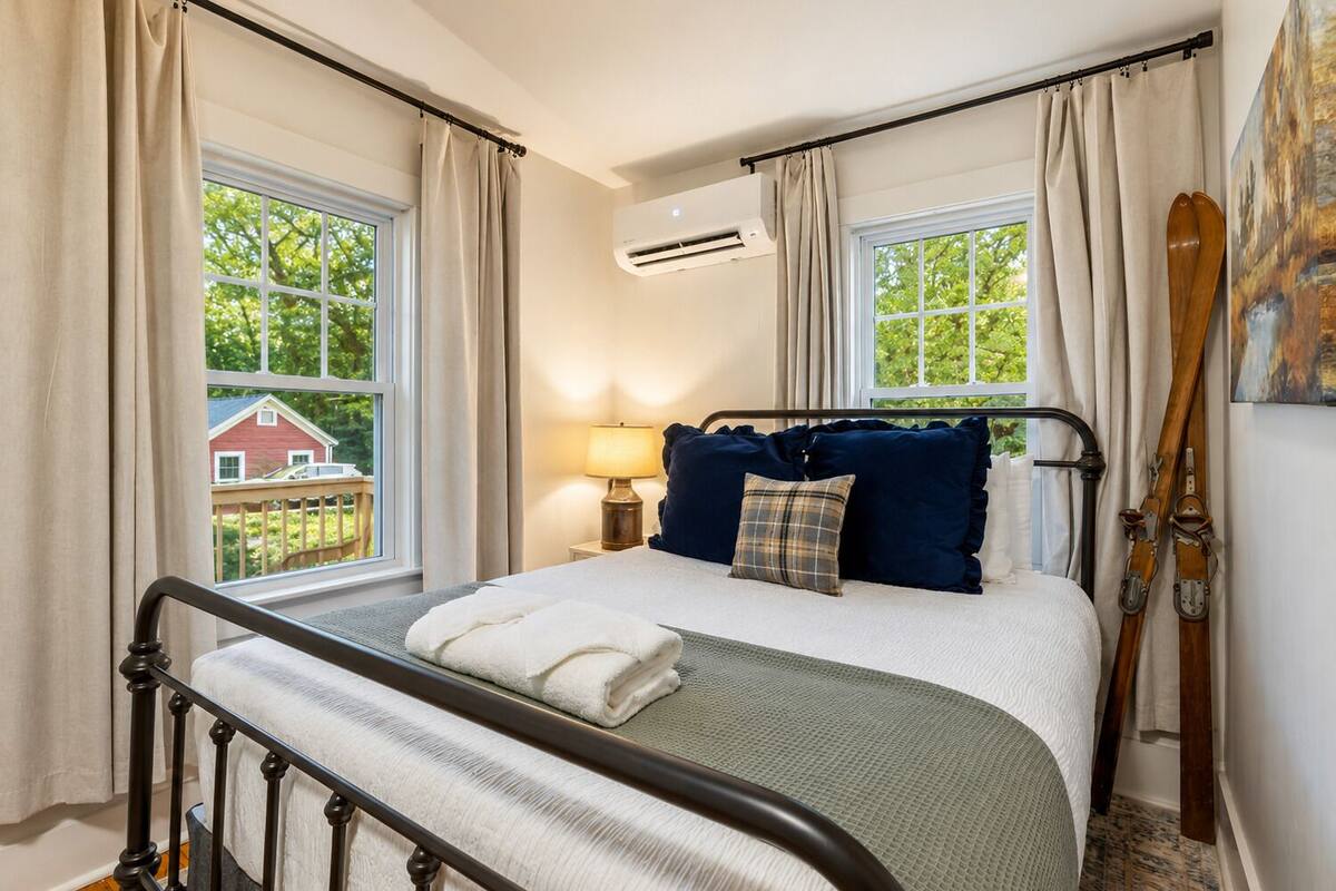 A bedroom with a bed, a window, an air conditioner, a lamp, and skis on the wall.