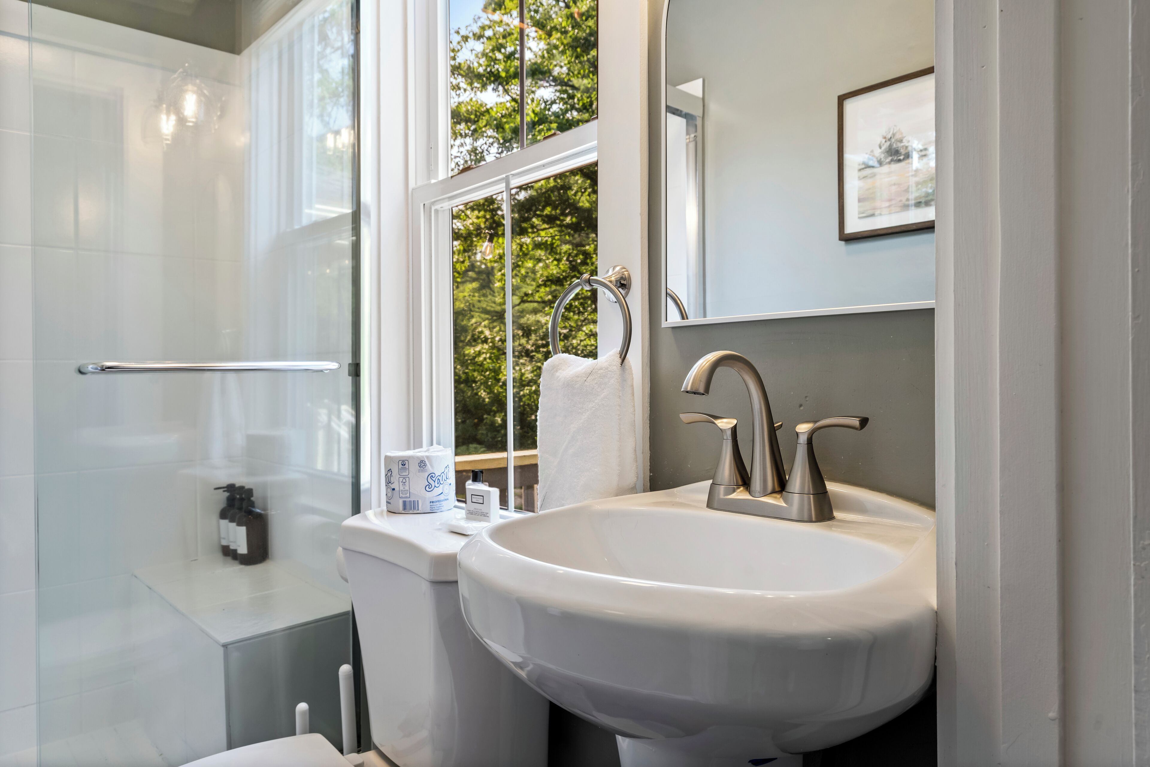 A bathroom with a glass shower enclosure, a white sink with a modern faucet, and a window with a towel hanging on the door.