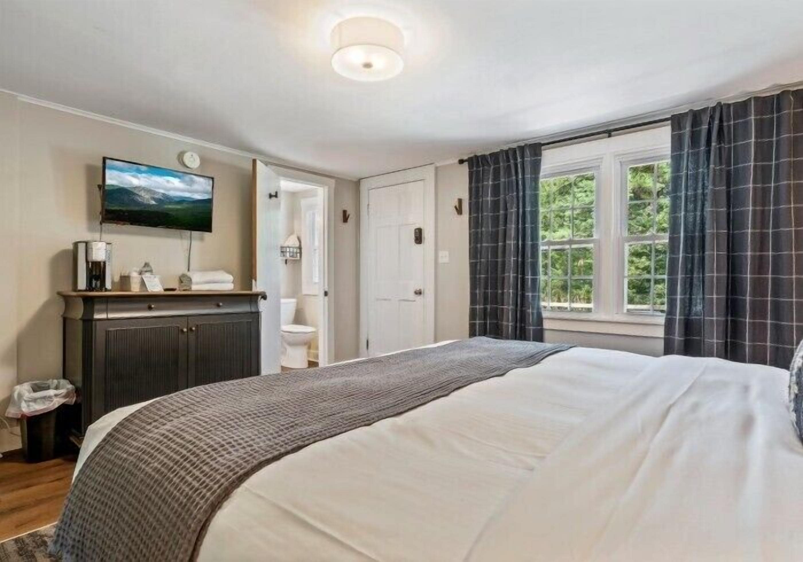 A bedroom with a bed, a television, a dresser, a window with curtains, and a door leading to a bathroom.