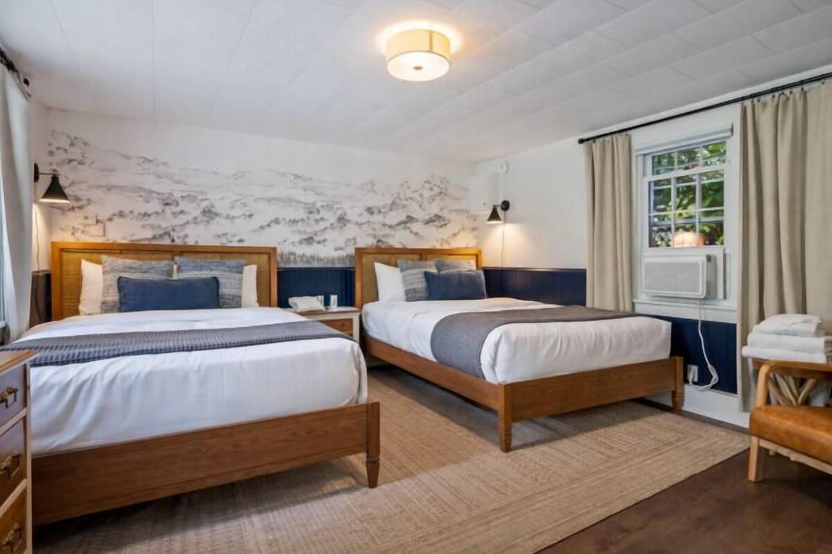 A double bed room with two beds, a wooden headboard, a bedside table, a lamp, a window with curtains, and a wall with a marble-like texture.