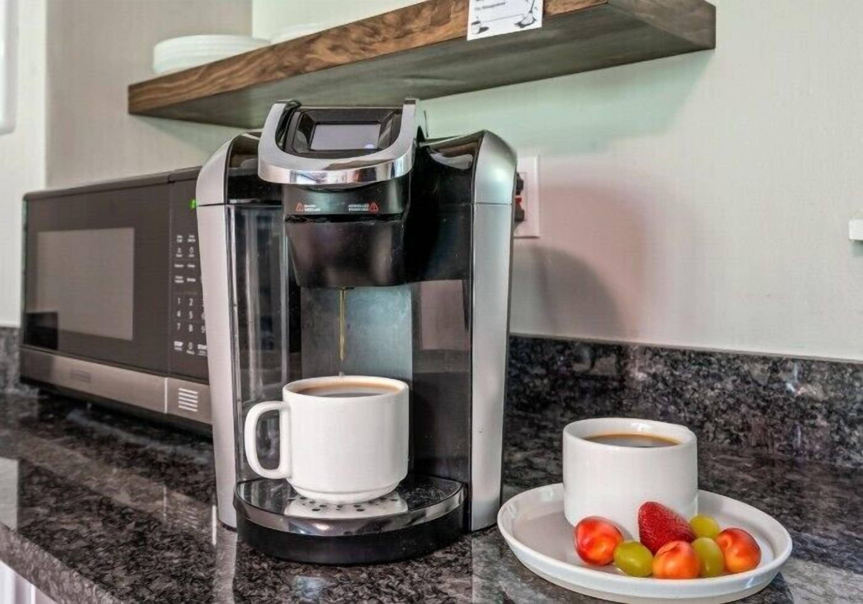 Une machine à café avec deux tasses, un four à micro-ondes et une assiette de fruits posés sur un plan de travail en granit.