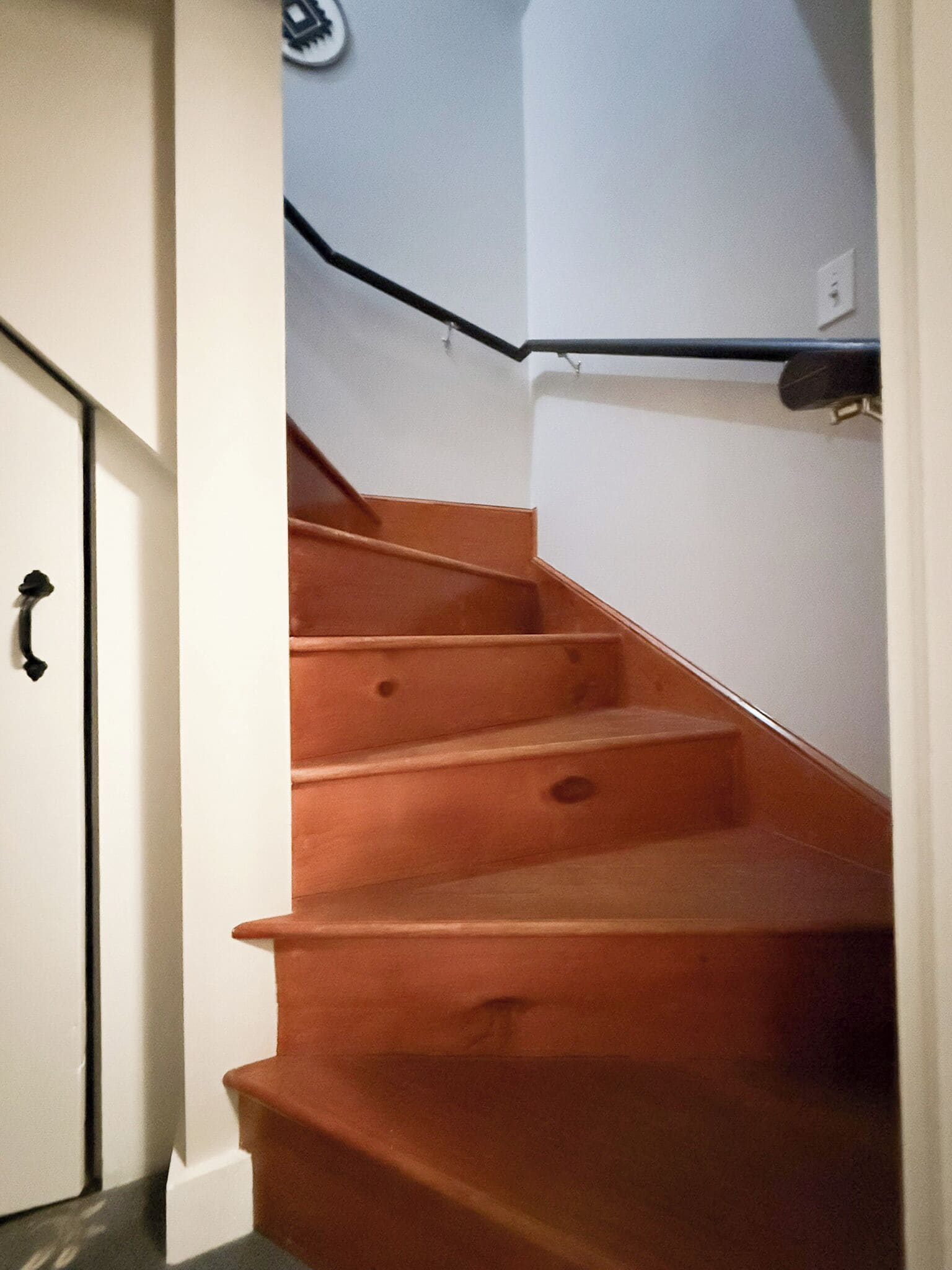 A wooden staircase with a handrail, white walls, and a door with a black handle.