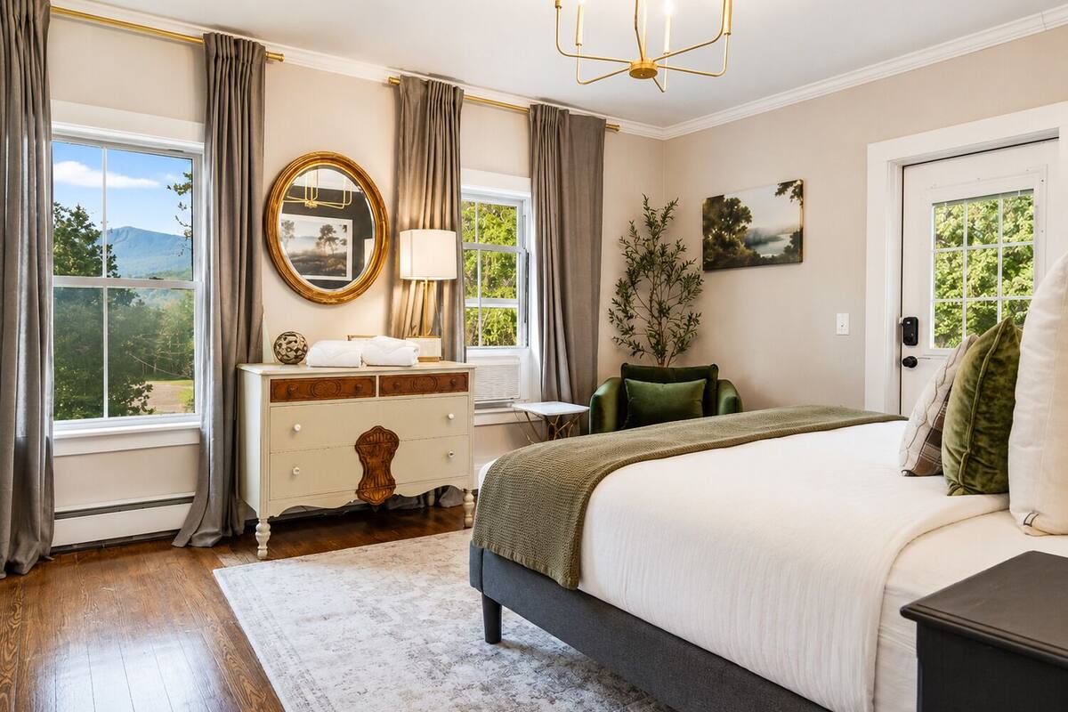 A bedroom with a large bed, a dresser, a mirror, a chair, and a view of the outdoors.
