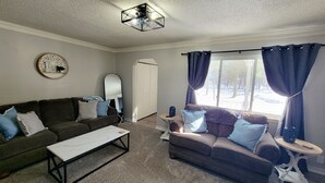 Ein Wohnzimmer mit einem dunkelbraunen Sofa, einem weißen Couchtisch und einem grauen Teppich. Ein Fenster mit blauen Vorhängen bietet Blick auf Bäume draußen.