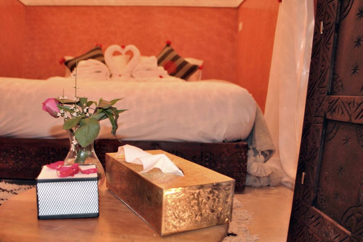 A neatly made bed with pillows and a heart-shaped decoration, a vase with flowers, and a tissue box on a wooden table.