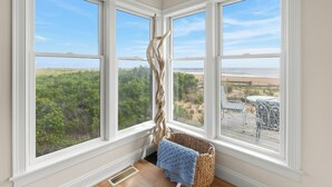 A corner room with three large windows, a wicker basket with a blue towel, and a view of a beach and greenery.