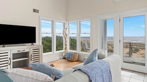 A living room with a large TV, a sofa, and a view of the beach.